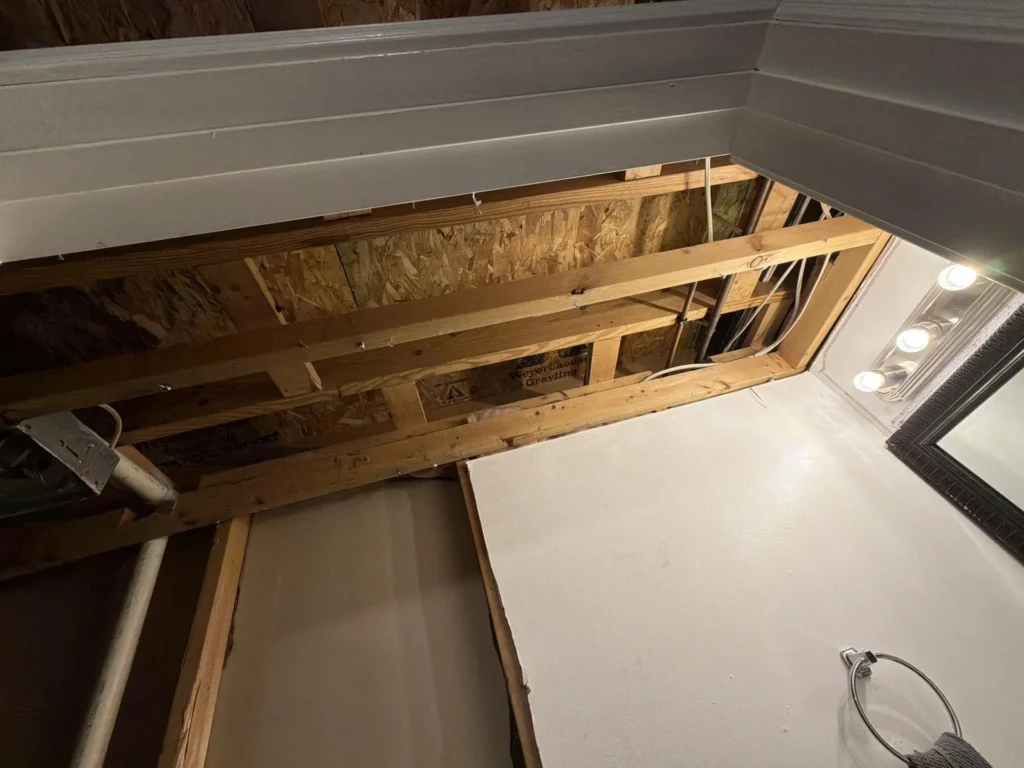 Bathroom with large section of ceiling removed exposing floor joists, OSB subfloor above, and wiring, indicating water damage investigation or repair, vanity mirror and light fixture still in place