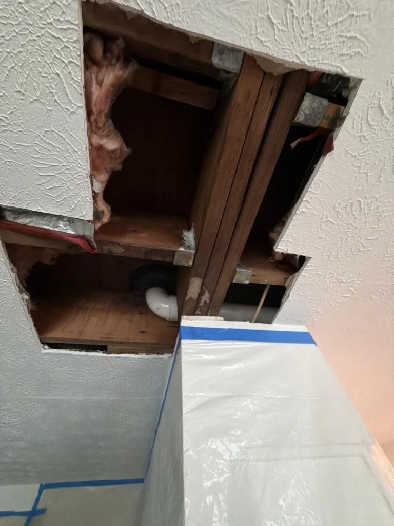 Ceiling of a room opened during water damage investigation showing wood joists, a PVC drain elbow, and aged insulation with plastic containment barrier taped with blue tape protecting surfaces below during remediation