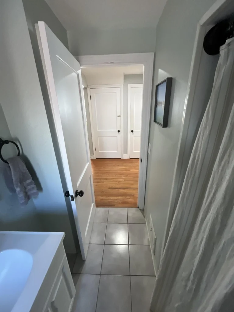 View through a bathroom doorway looking into a hallway with hardwood floors, showing white doors, tile flooring inside the bathroom, a shower curtain, and white vanity