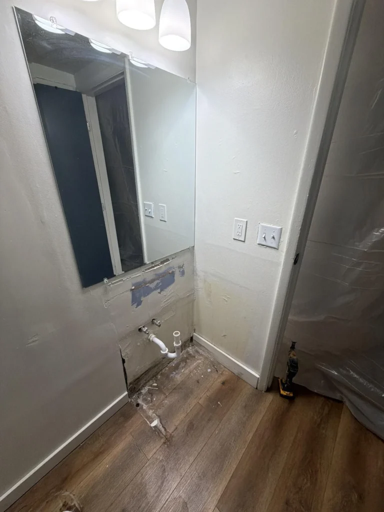 Bathroom vanity area with sink removed, lower wall drywall cut away showing PVC supply lines and P-trap stub-out, water-damaged hardwood floor with adhesive residue, mirror and vanity light still in place