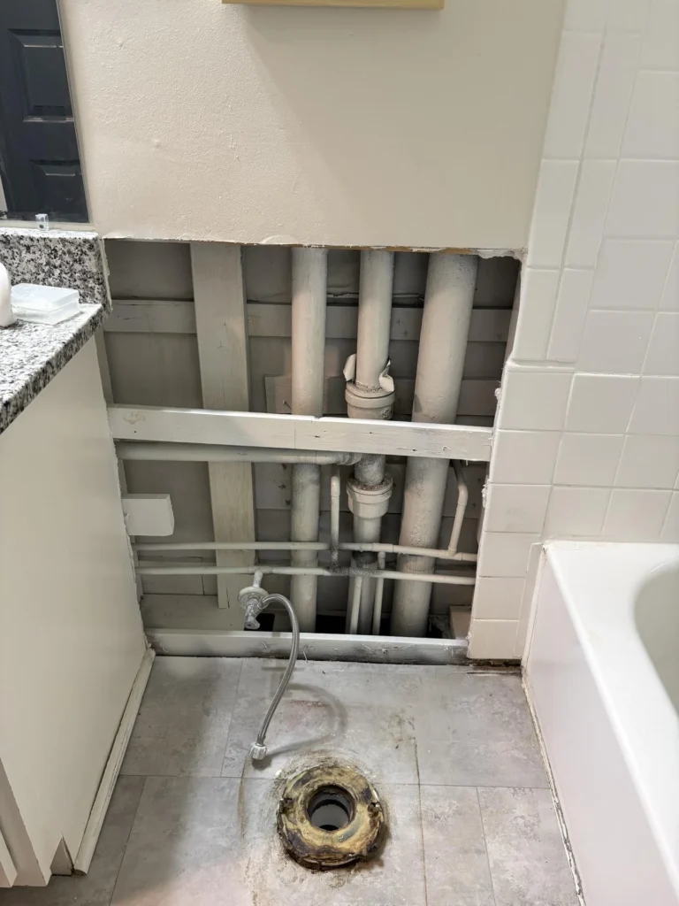 Bathroom with wall drywall cut open from floor to mid-height exposing multiple PVC drain pipes and water supply lines, toilet removed leaving exposed floor flange, granite vanity top and white tile tub surround still intact