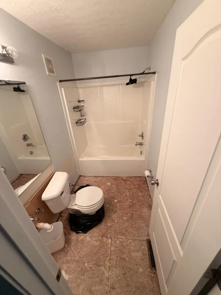 Overhead view of a bathroom during water damage assessment with floor tile removed exposing damaged OSB subfloor, white bathtub with tile surround and shower rod, toilet with garbage bag, vanity with exposed plumbing, and textured ceiling