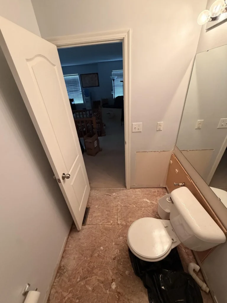 Water-damaged bathroom with floor tile removed showing damaged OSB subfloor, open door to a carpeted bedroom with boxes visible, toilet, exposed vanity cabinet framing, and vanity light with mirror