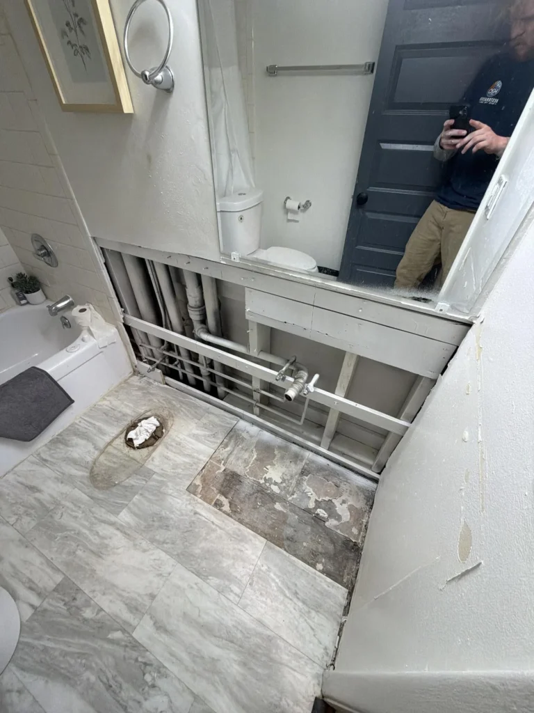 Bathroom with lower wall section cut open exposing multiple supply and drain pipes in a mirror reflection, toilet flange stub-out on floor, water-damaged tile beneath vanity area, bathtub and towel bar still in place