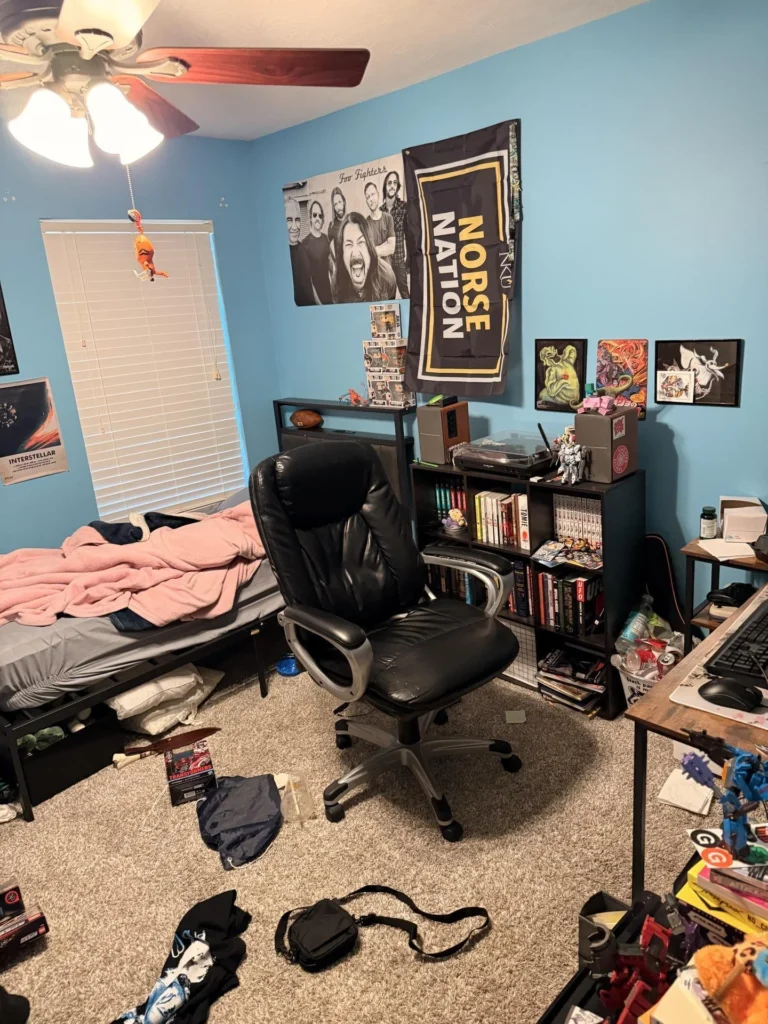 Cluttered bedroom with blue walls, black leather office chair, bed with pink blanket, bookshelf with manga and books, ceiling fan with light, window with blinds, and various band and anime posters including Foo Fighters and Norse Nation flag