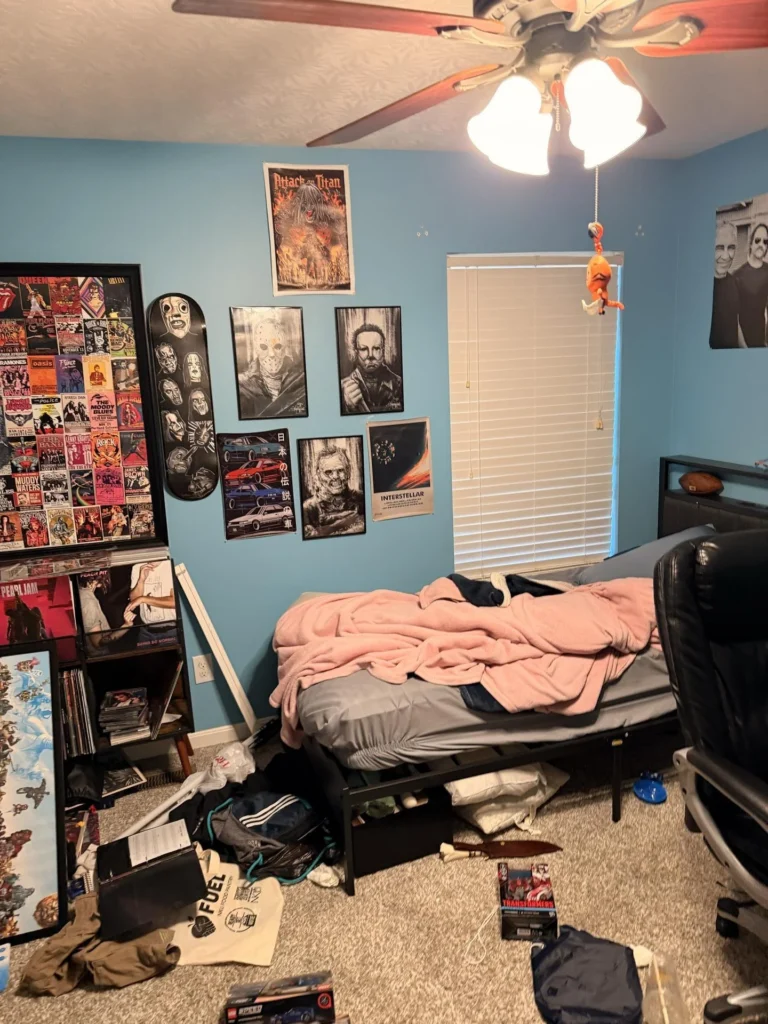 Bedroom with blue walls covered in horror, anime, and pop culture posters and artwork, vinyl record collection and bookshelf on the left, unmade bed with pink blanket, ceiling fan, window with blinds, and various items on the floor