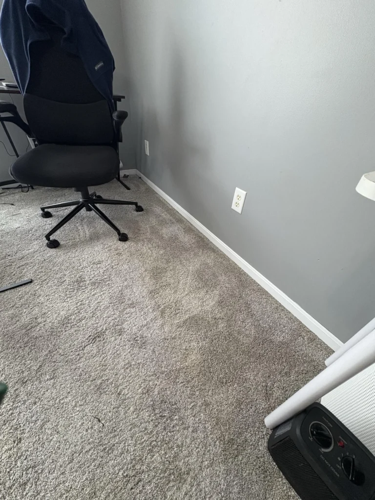 Bedroom with gray carpet showing water damage staining near the baseboard, a black gaming chair, gray walls, and a portable heater on the floor during a water damage assessment