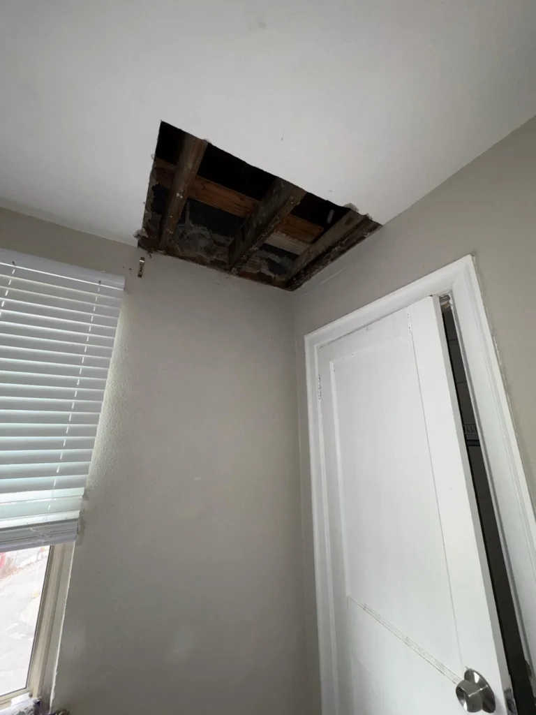 Bedroom corner with a large rectangular section of ceiling drywall removed exposing dark water-stained wood joists and plank subfloor above, beside a window with blinds and an open closet door with water-stained frame