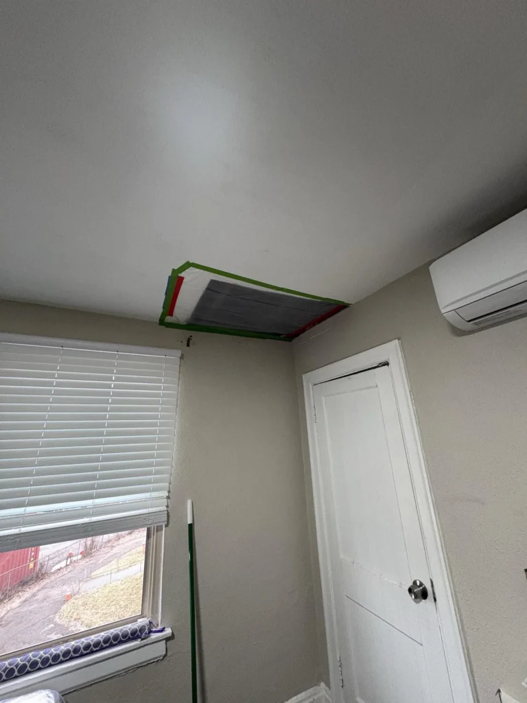 Bedroom ceiling after partial repair showing a dark gray plastic sheet sealed with green, red, and blue painter tape covering the repaired drywall opening, with a window, closet door, and wall-mounted mini-split unit visible