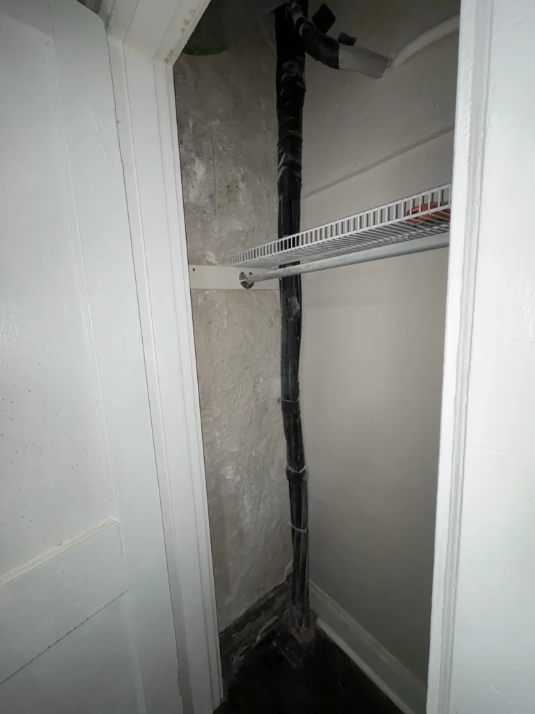 Interior of a narrow bedroom closet showing a heavily mold-stained and water-damaged back wall with a large black insulated refrigerant line running vertically, a white wire closet shelf and hanging rod, and a damaged white door frame