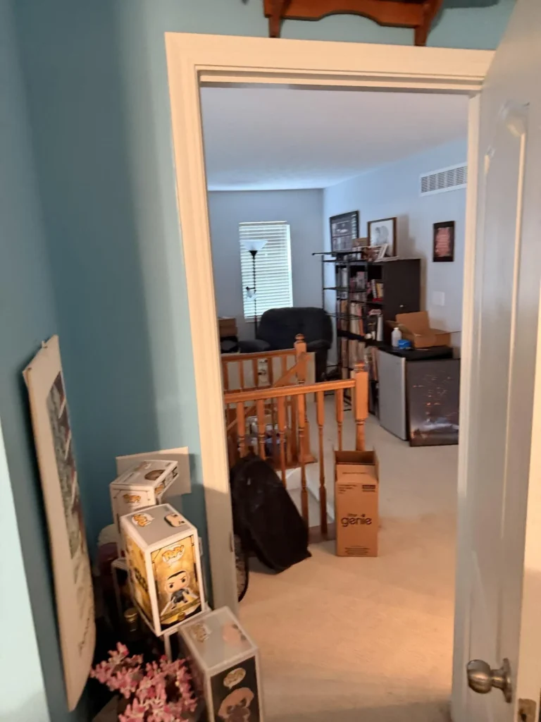 View from blue-walled bedroom doorway looking into upstairs hallway showing wood stair railing, carpeted floor, shelving with books and media, and collection of boxed Funko Pop figures stacked by the door frame