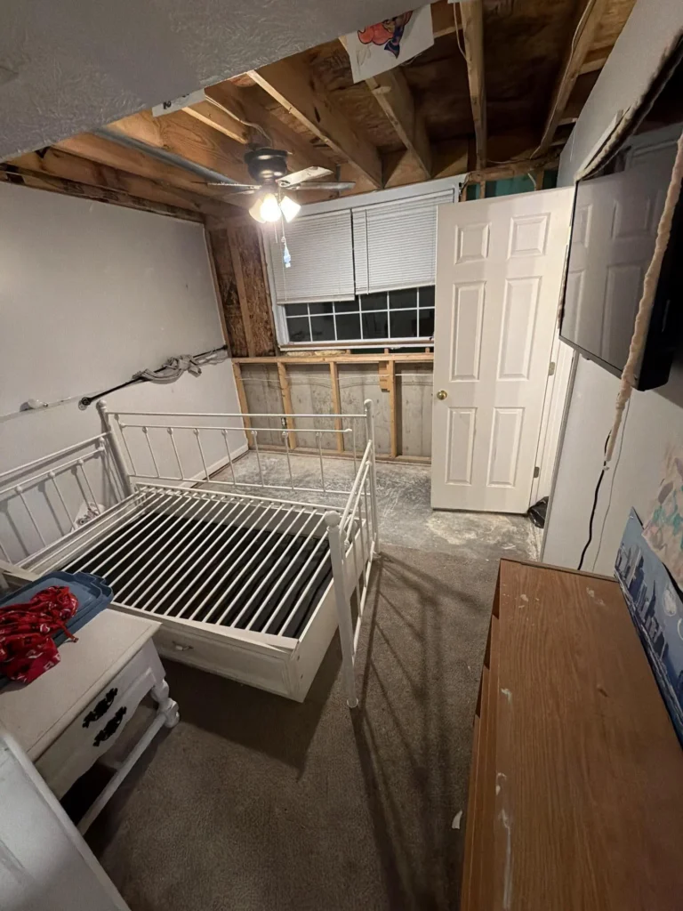 Child bedroom with large section of ceiling and wall drywall removed exposing wooden framing and OSB sheathing, white metal bed frame without mattress, dresser, and window with blinds intact