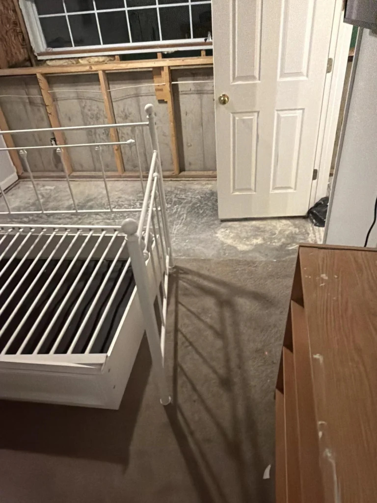 Flood-damaged small bedroom showing lower wall drywall removed exposing bare wood studs and sheathing, white metal bed frame, closed door, and dusty bare concrete floor suggesting significant water damage