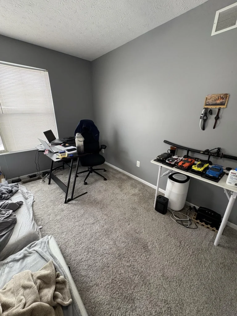 Bedroom with gray walls, gray carpet, a gaming desk setup with laptop and gaming chair, a side table with model cars, and a portable air purifier during a water damage inspection