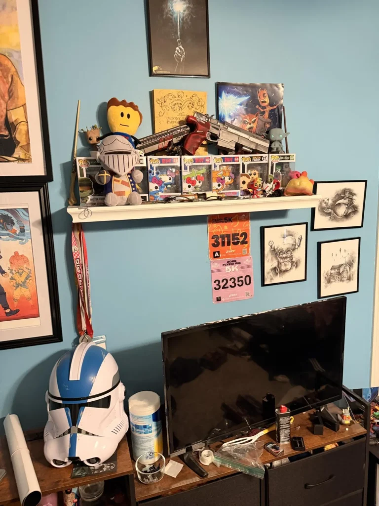 Bedroom display shelf on blue wall featuring Funko Pop vinyl figures, a Star Wars clone trooper helmet, toy guns, plush toys, race bib numbers, framed artwork, and a flat-screen TV below, with more framed art on surrounding walls