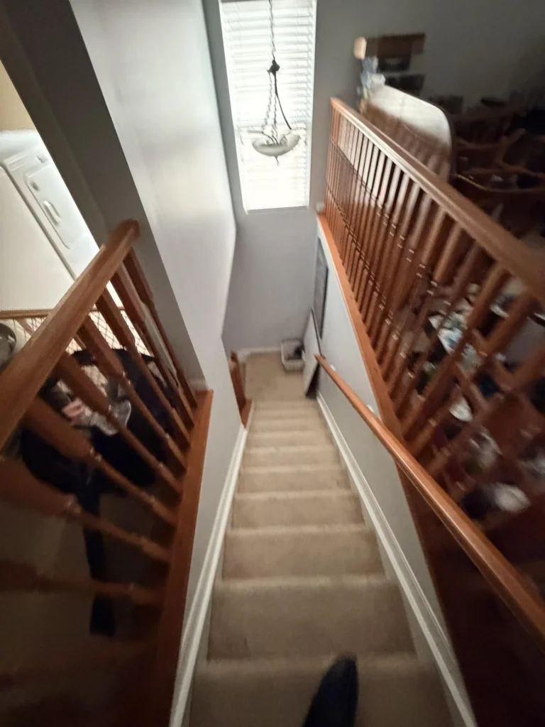 Looking down a carpeted staircase with oak wood baluster railing on both sides, pendant chandelier visible through stair window, washer on the left, and cluttered loft area with furniture visible from above