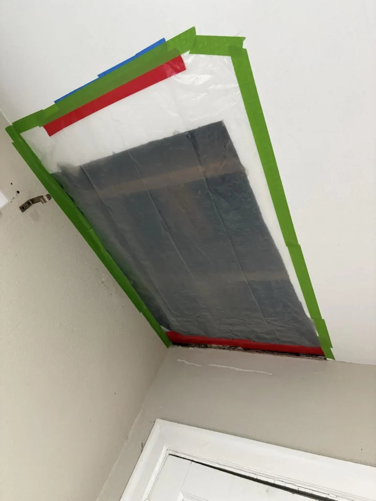 Bedroom ceiling with a large section covered in layered plastic sheeting sealed with green, red, and blue painter tape to contain an active water leak, with a damaged white door frame visible below