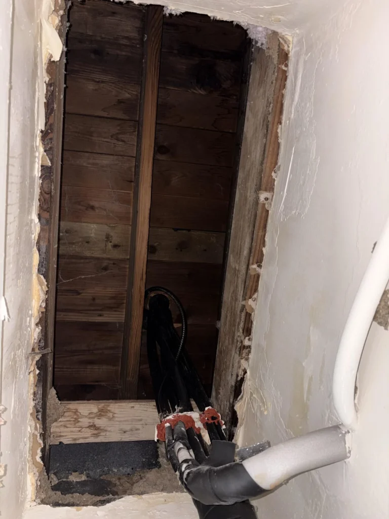 Close-up of a rectangular access hole cut through drywall inside a closet revealing dark stained wood subfloor framing and wall cavity, with black insulated refrigerant lines and red pipe fittings visible at the opening