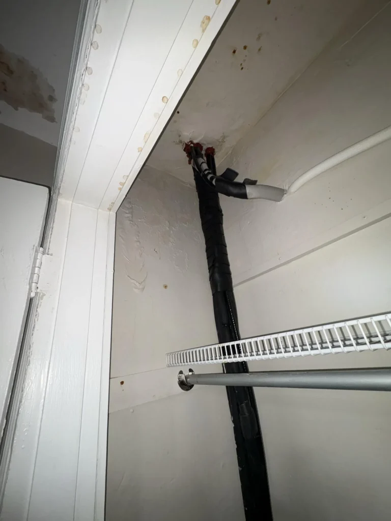 View inside a bedroom closet looking up at the ceiling and door frame header showing numerous brown water stain spots and mold around where a black insulated mini-split line and white pipe pass through, with a wire shelf and closet rod visible