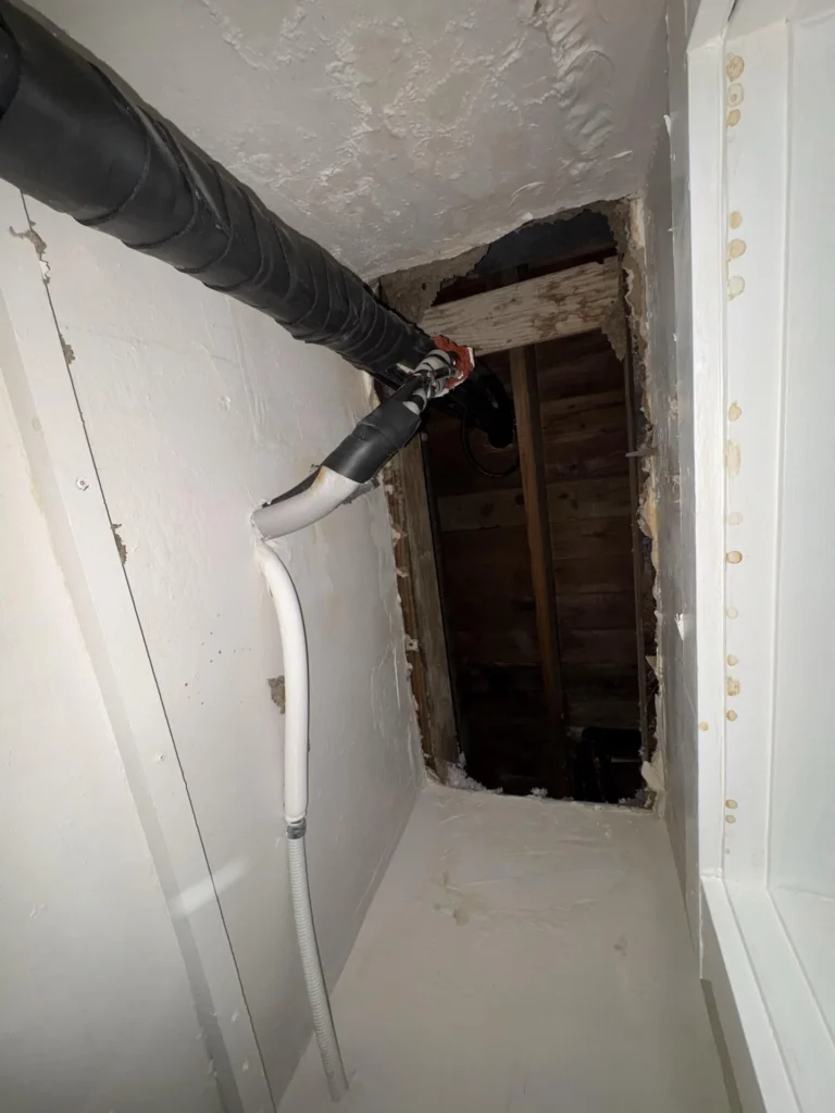 Inside a closet showing a rectangular access hole cut through the wall exposing dark water-stained wood framing behind, with a large black insulated mini-split line and white flexible condensate hose routed through the opening