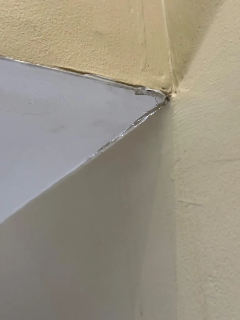 Close-up of a ceiling corner showing separated and cracking drywall joint tape along the seam where white ceiling drywall meets tan-painted walls, with lifted compound edges and darkened moisture damage at the joint
