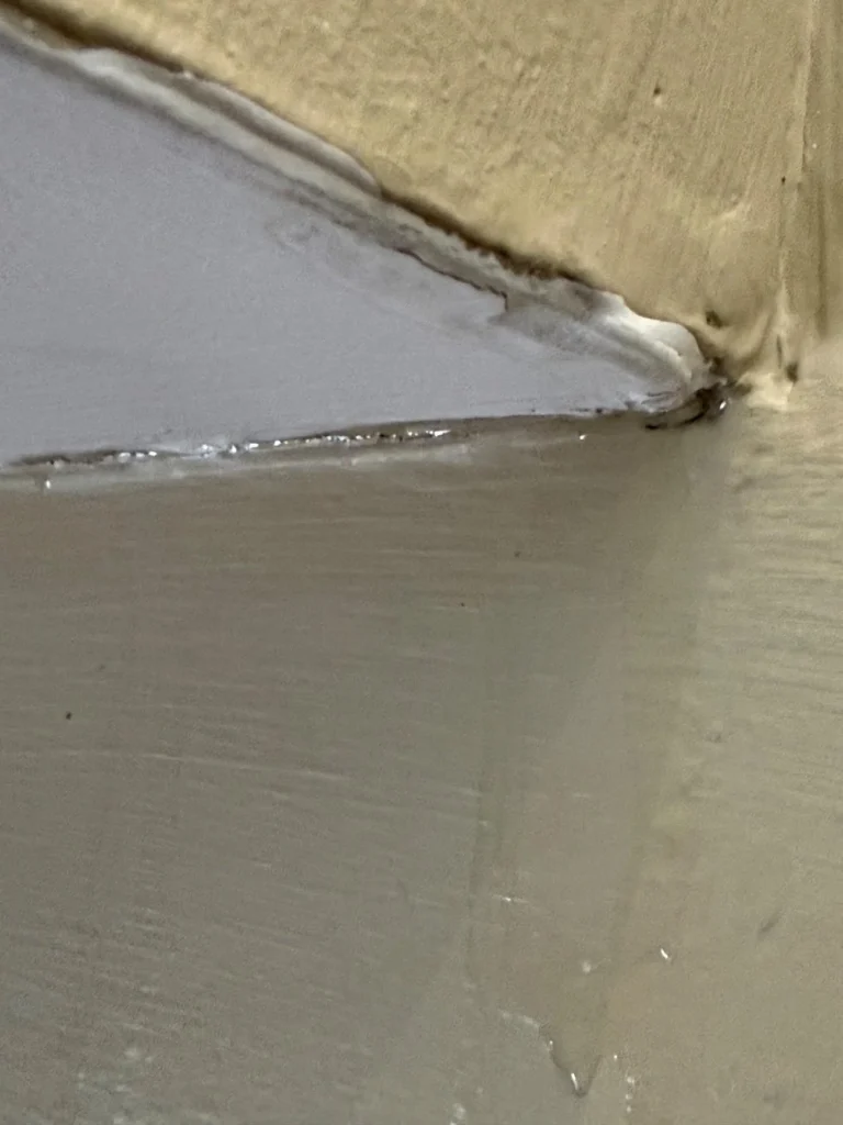 Extreme close-up of a ceiling corner joint where drywall tape and joint compound have separated and cracked along the seam between a white ceiling and tan-painted walls, with peeling and moisture damage visible