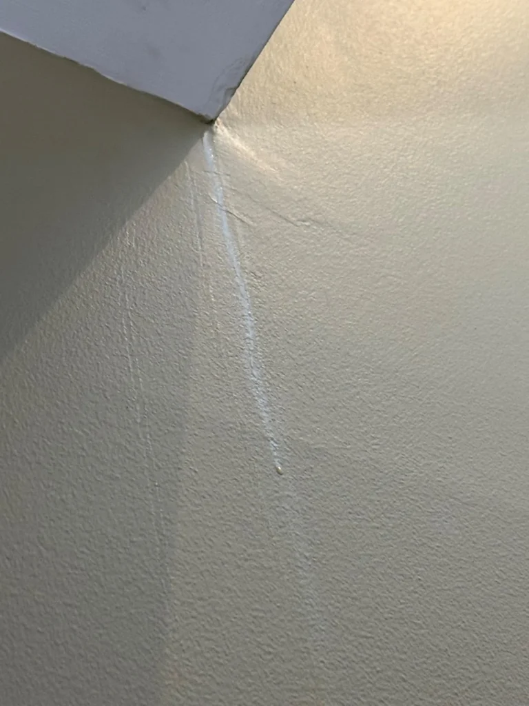 Close-up of a ceiling-to-wall corner showing a long diagonal crack in the drywall joint tape that has separated and lifted away from the surface, with a pale yellow wall texture visible in the upper corner