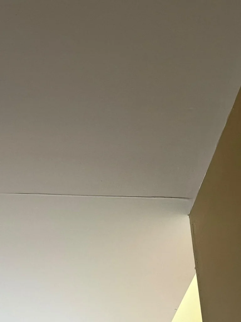 Corner of a room showing the ceiling meeting a white wall on the left and a tan wall on the right, with a visible seam separation or crack running along the ceiling drywall joint line