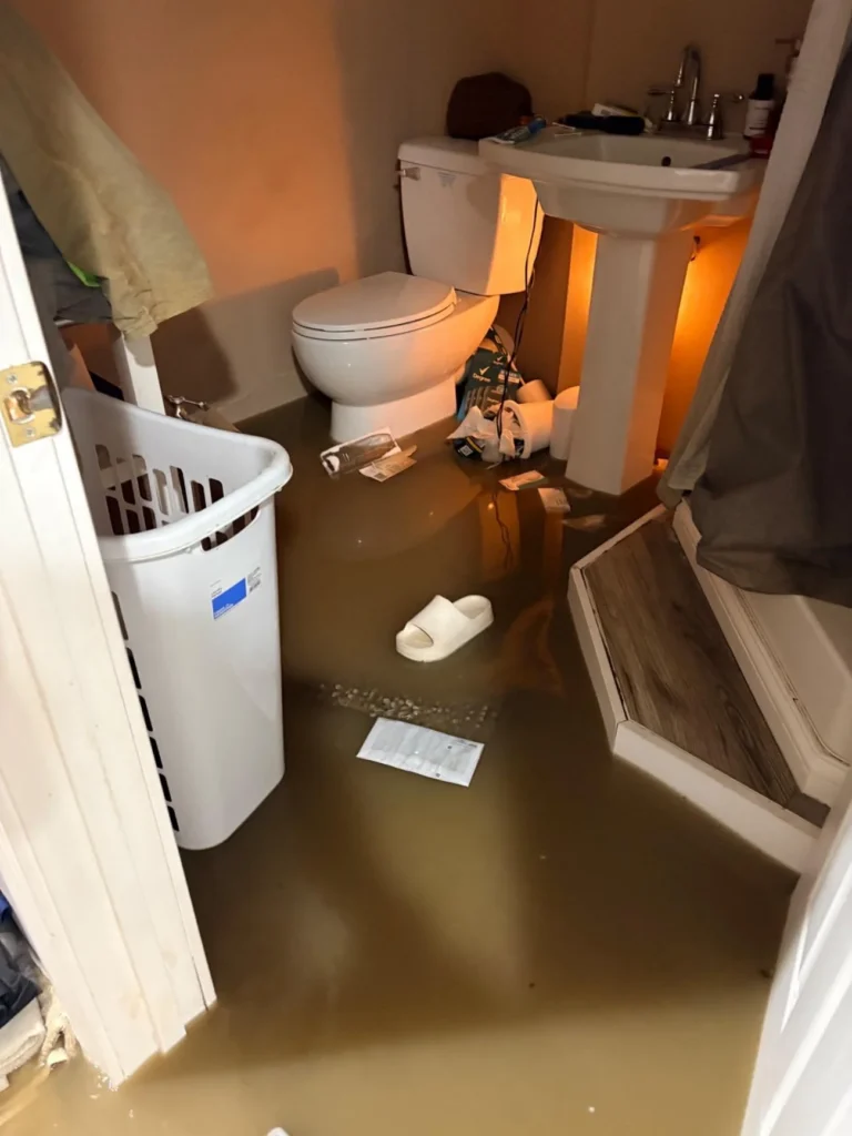 Flooded basement bathroom with brown water covering the tile floor, a toilet, pedestal sink, laundry basket, floating slipper and paper, and a step stool, all submerged in standing water