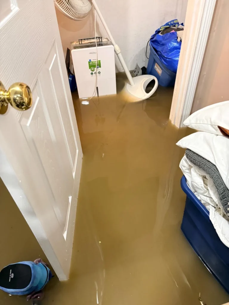Flooded basement hallway with murky brown water covering the floor, a Soleus Air 45-pint dehumidifier running in the corner, blue storage bins, a fan, and a mop tip visible amid standing water