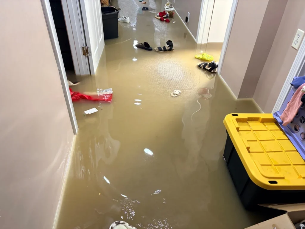 Long flooded basement hallway with brown standing water, multiple shoes floating including sandals and crocs, open doorways to adjacent rooms, a yellow-lidded storage bin, and scattered debris throughout