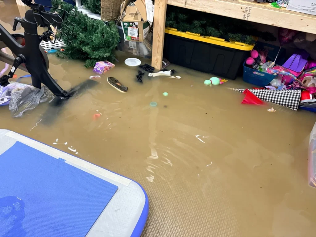 Flooded basement with deep brown standing water, a Peloton bike, children's toys and shoes floating, a wooden storage shelf with totes and holiday decorations, and a blue yoga mat in the foreground
