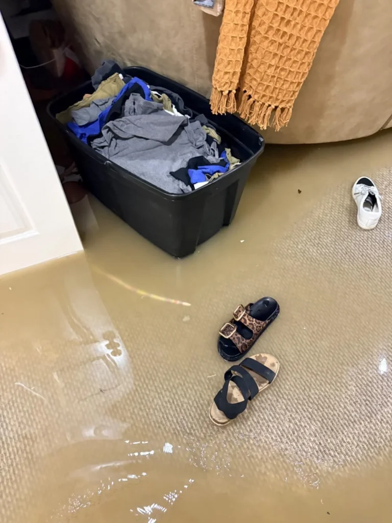 Flooded basement room with brown water on the carpet, two pairs of sandals floating, a black storage bin full of clothes, a beige sofa with an orange throw, and a white sneaker drifting in the water