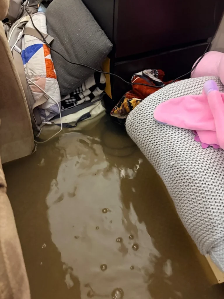 Close-up of brown floodwater on a basement bedroom floor between a sofa and dark dresser, with soaked clothing, charging cables, a stuffed animal, and a pink toy visible in the standing water