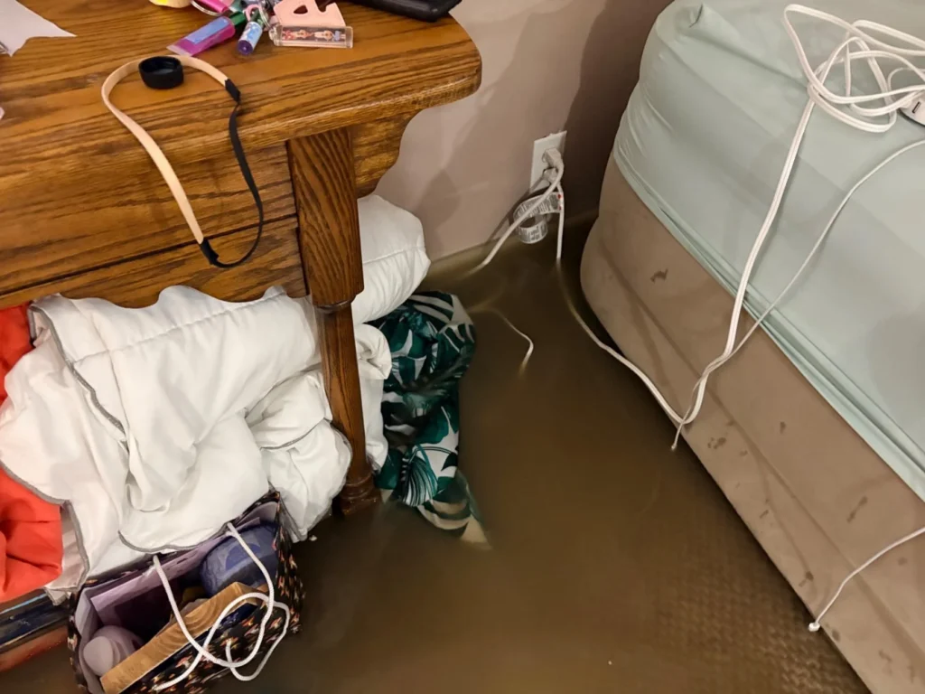 Flooded basement bedroom with brown water covering the carpet, a wooden nightstand with personal items, a soaked white comforter and tropical-print blanket submerged under the bed near a flooded electrical outlet