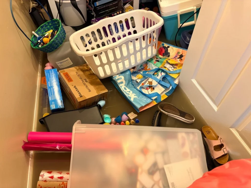 Flooded basement closet or utility room with murky water, an overturned laundry basket, cardboard boxes, scattered art supplies, shoes, a tote bag, and a clear plastic storage bin amid the standing water