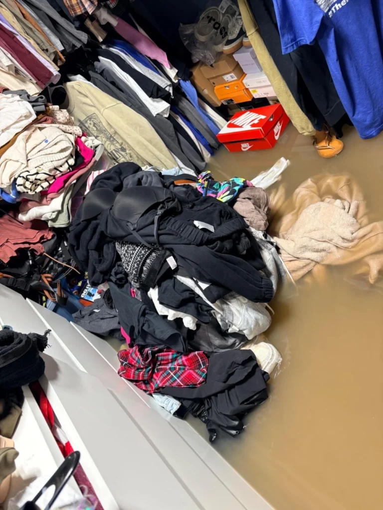Flooded walk-in closet with brown standing water, piles of wet clothing fallen from hangers on the floor, Nike and other shoeboxes getting soaked, and clothes racks visible in the background