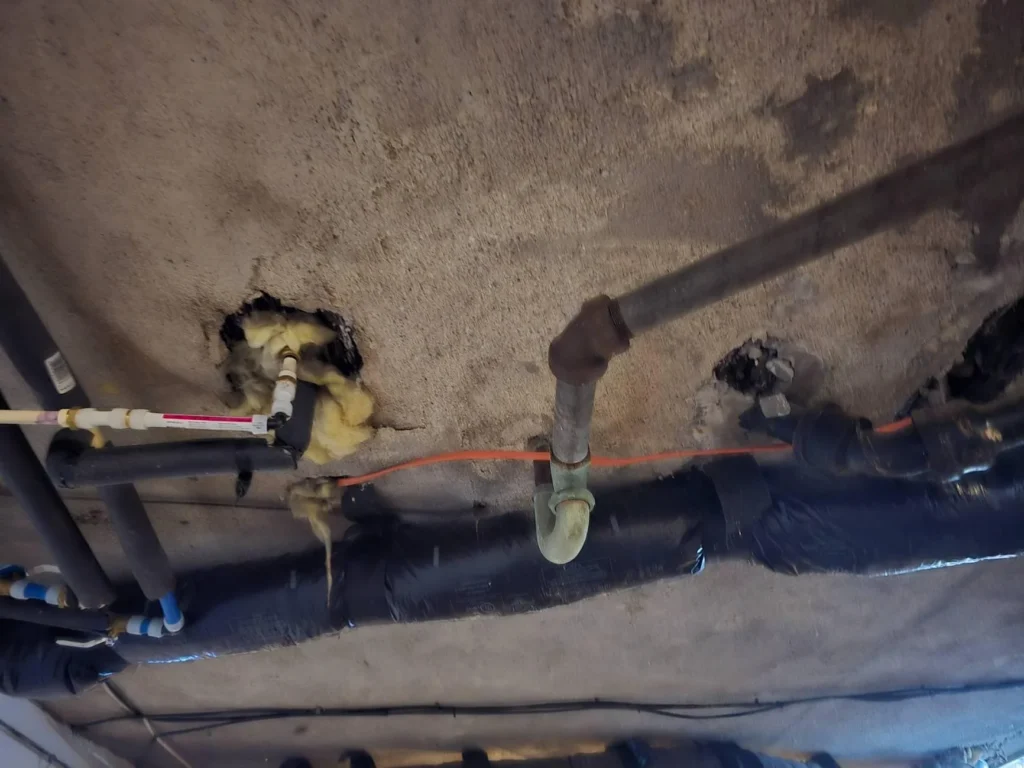 Two workers inspecting a garage or basement space with white-painted cinder block walls, a concrete floor with leaf debris and stains, and a damaged ceiling exposing black ductwork and pipes