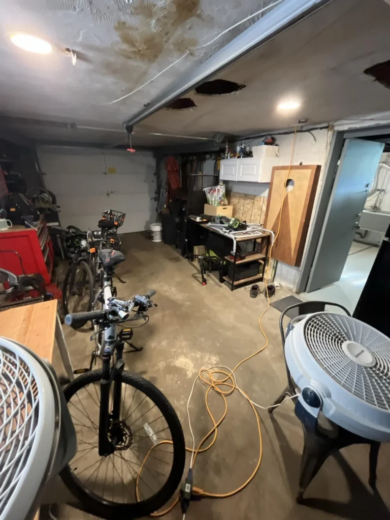 Garage interior viewed from the front showing severely water-stained ceiling with two cut-out inspection holes, bicycles parked inside, and large floor fans running for drying