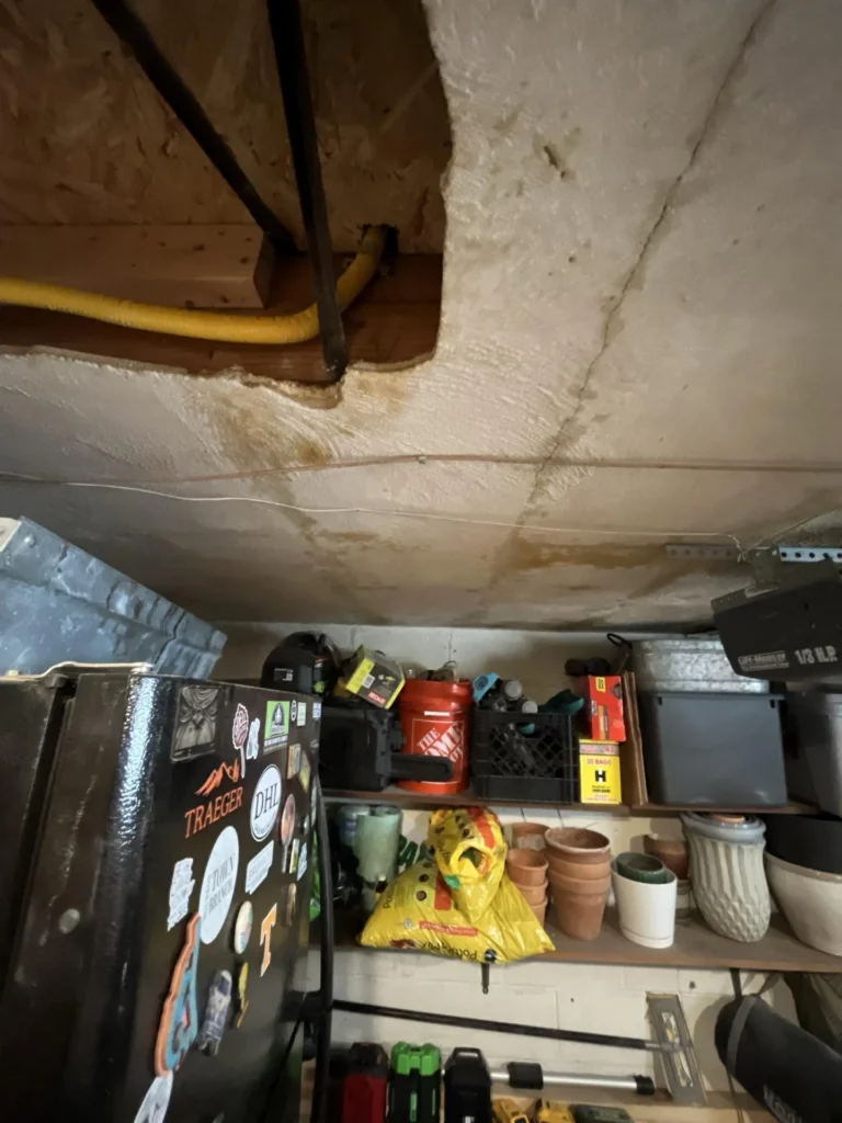 Close-up of garage ceiling showing brown water staining and damage near an exposed gas line access panel, with shelving holding gas cans, terra cotta pots, and storage containers below