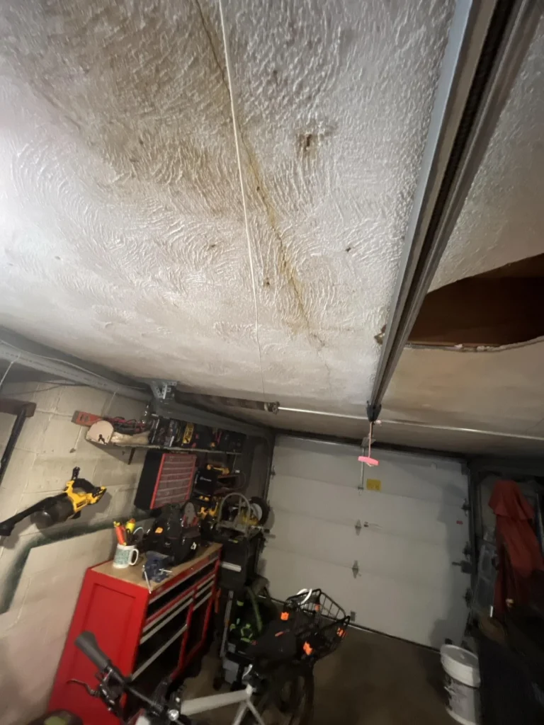 Garage ceiling with extensive brown water damage stains and discoloration, showing a large affected area above a workshop with tool chests, power tools, and bicycles below