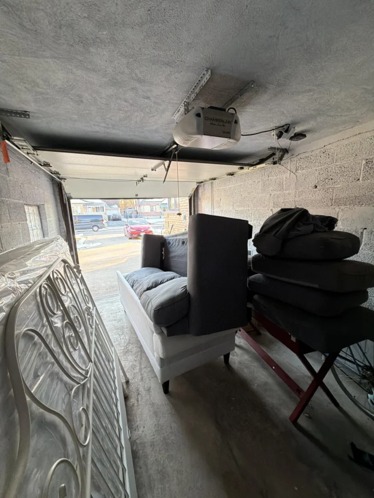 Garage interior with door partially open and a residential street visible outside, storing a gray upholstered chair, stacked dark sofa cushions on a folding stand, and a white ornate metal headboard, with a Chamberlain opener overhead