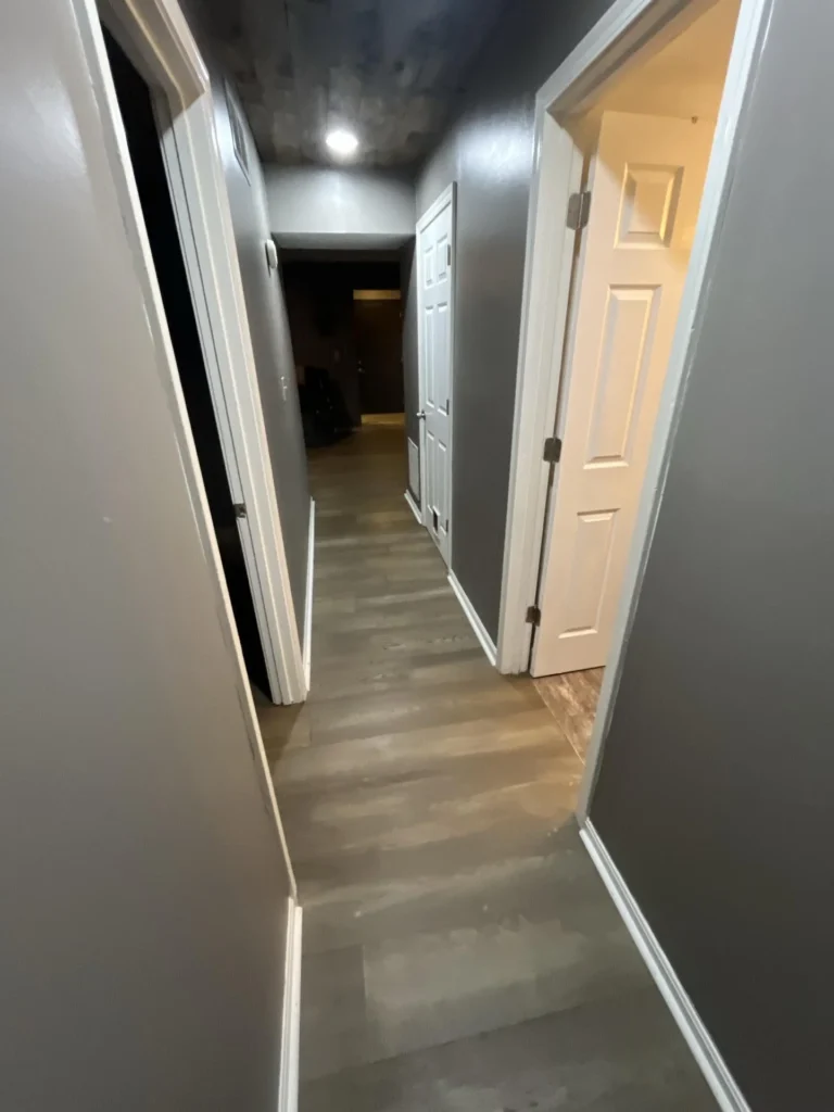 View down a residential hallway with gray walls, wood-look LVP flooring, white trim, and an open door revealing a lit room at the end during a water damage inspection