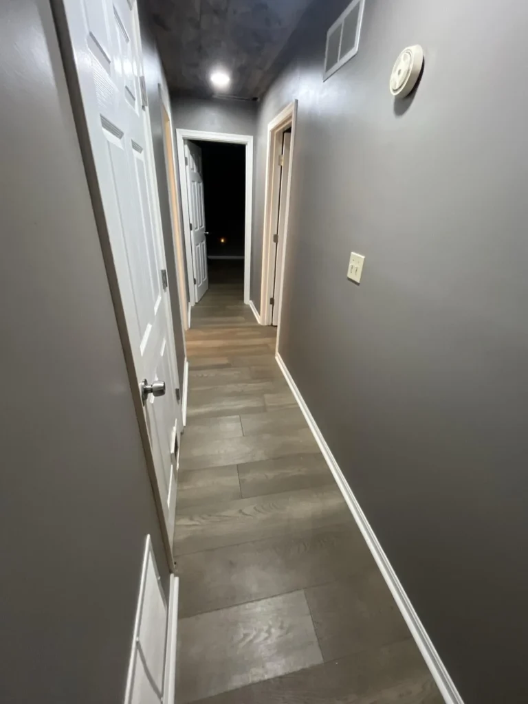 Alternate angle of a residential hallway with gray walls, wood-look LVP flooring, white door trim, light switch plate, and a smoke detector on the wall during water damage remediation