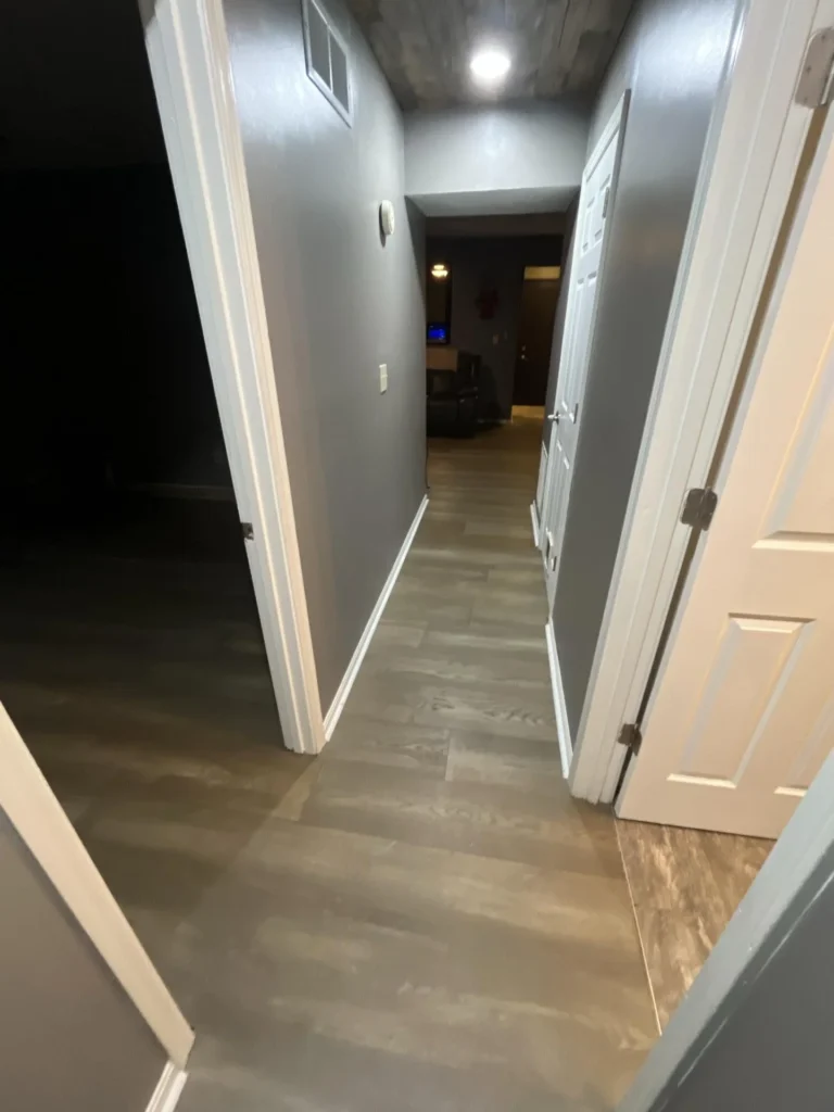 Narrow residential hallway with gray walls, luxury vinyl plank flooring, multiple white panel doors, and a recessed ceiling light showing signs of water damage during a restoration assessment