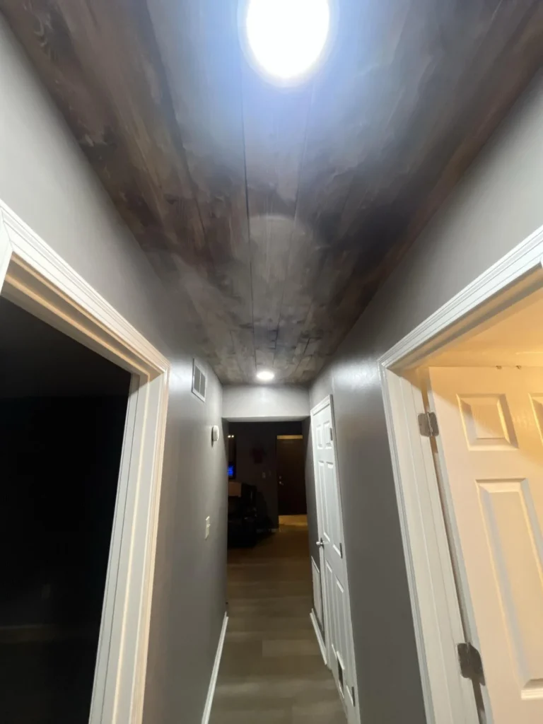 Residential hallway viewed from below showing a dark-stained wood plank ceiling with water damage discoloration, multiple white doors, and recessed lighting indicating moisture intrusion from above