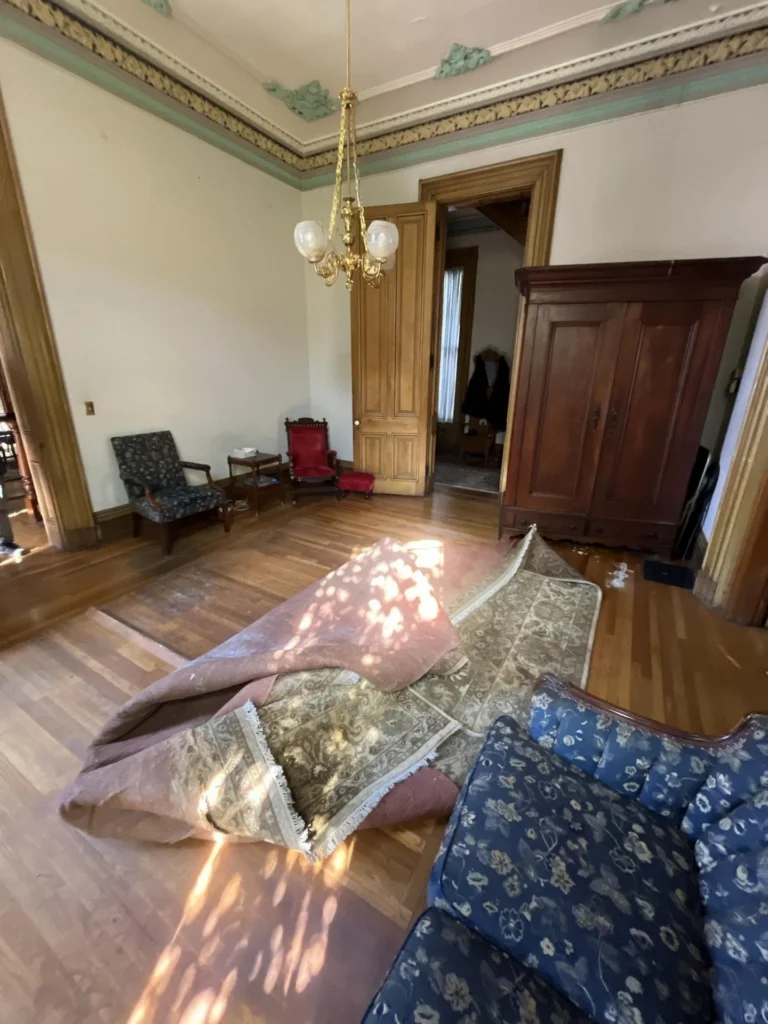 Elegant parlor of a historic home with ornate green and cream plaster crown molding, gold chandelier, tall wood-trimmed doorway, a large antique wardrobe, antique chairs, rolled-up rugs on hardwood floor indicating water damage assessment