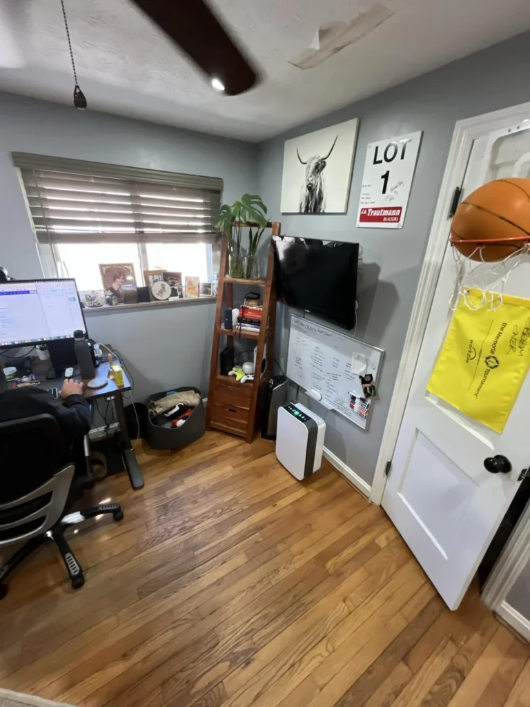 Home office room with hardwood floors and gray walls showing a water-damaged ceiling patch above a ceiling fan, with a computer workstation, wall-mounted TV, and whiteboard visible