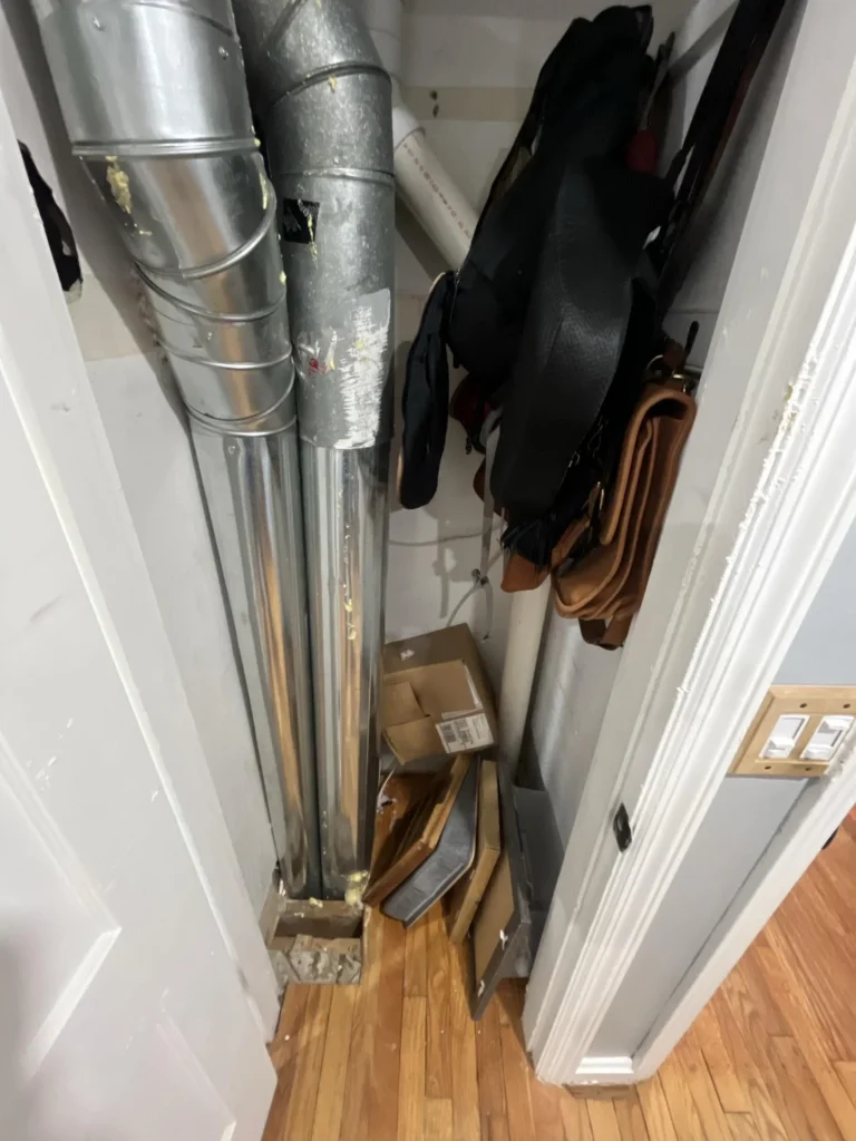 Closet interior with large galvanized steel HVAC ducts running floor to ceiling alongside damaged drywall, with handbags and boxes stored below and hardwood hallway floor visible