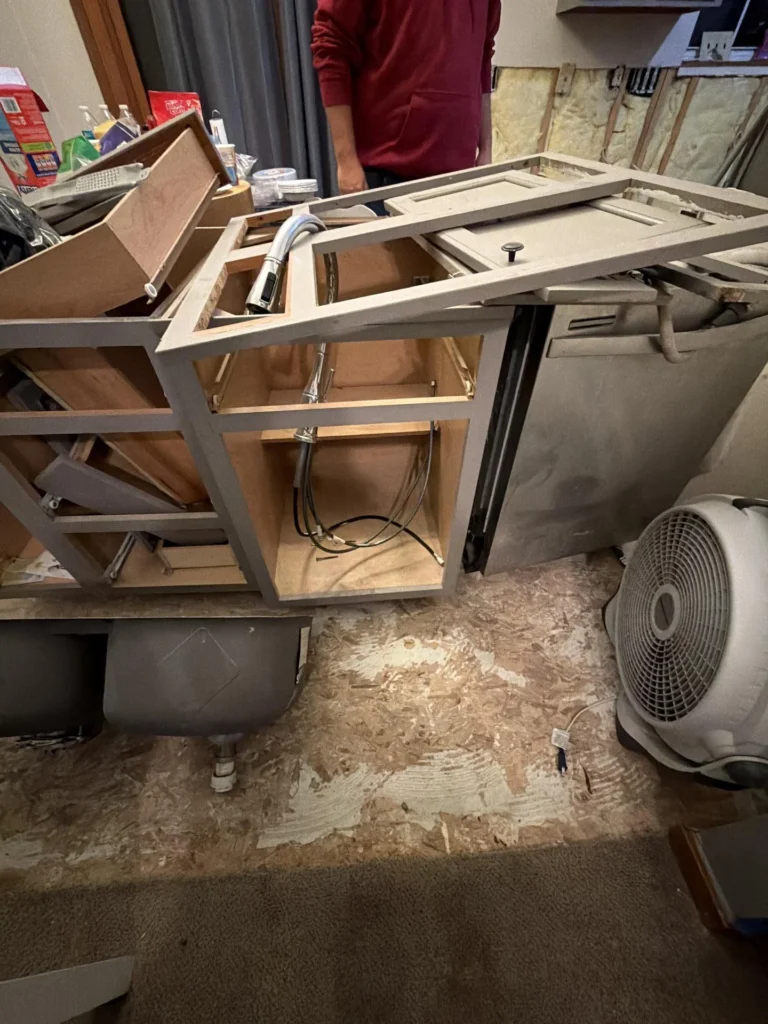 Flood-damaged kitchen with gray base cabinets removed and tipped over showing faucet and plumbing lines still attached, dishwasher pulled out, box fan on bare subfloor, exposed wall insulation in background